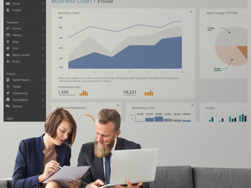Muškarac i žena sa laptopom i dokumentima analiziraju podatke, iza njih je dashboard sa grafikonima